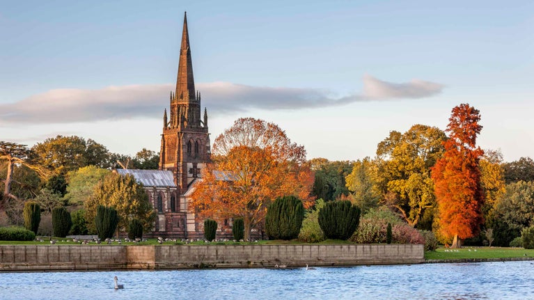 Across the lake, the tall spire of the chapel at Clumber Park, Nottinghamshire, is visible through trees which have turned the orange and golden colours of autumn.